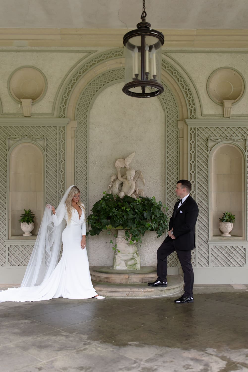 Bride and groom positioned beneath a stone fountain in a covered courtyard, framed by soft architectural details.
