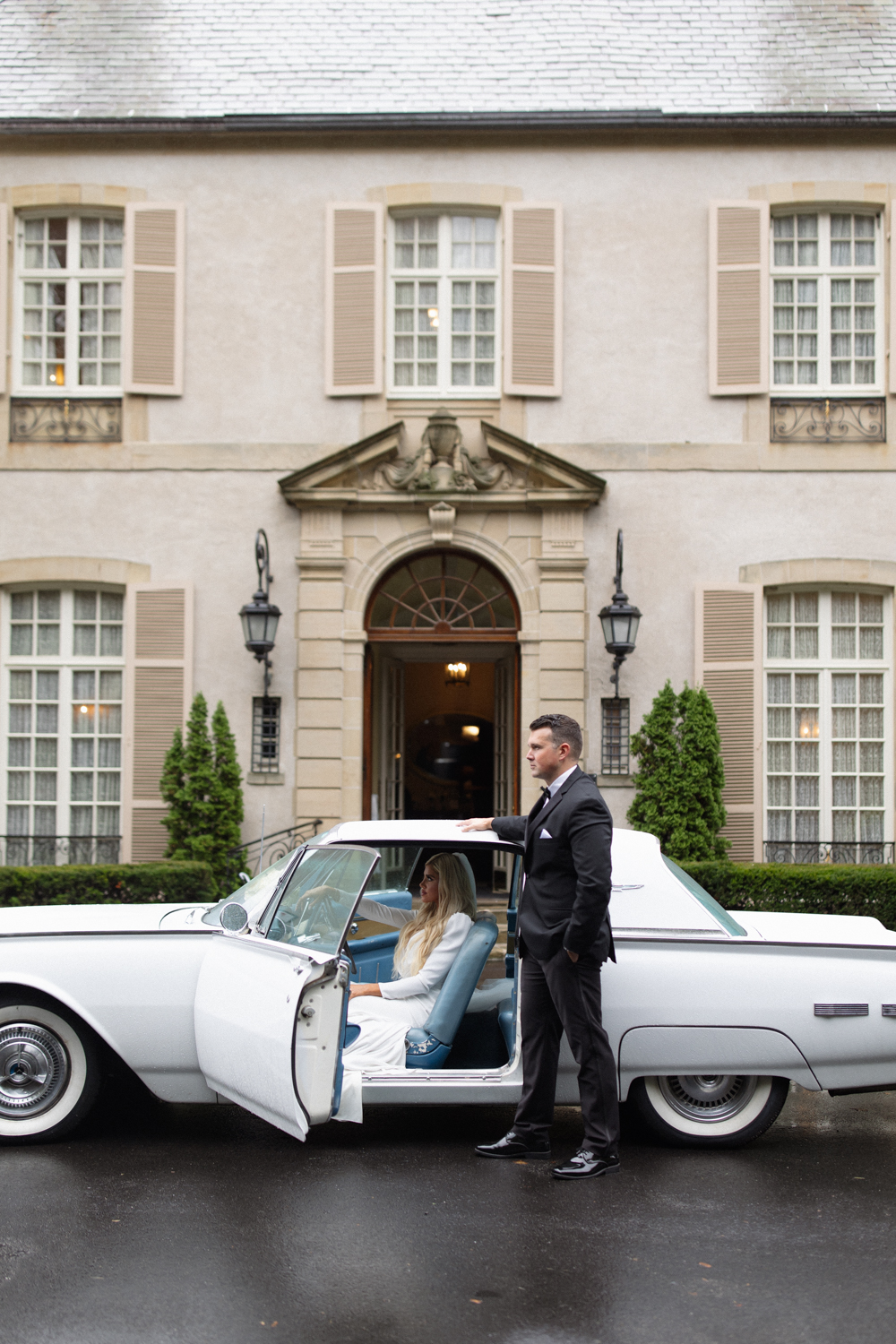Bride seated in a vintage white car as groom stands beside the open door in front of a historic estate wedding venue in Rhode Island.
