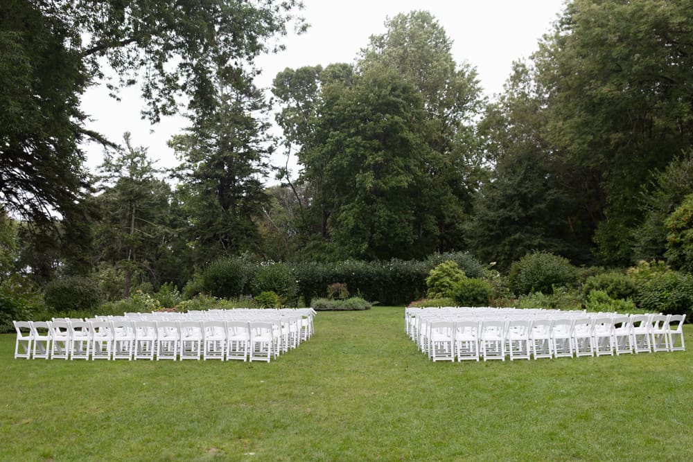 Outdoor ceremony chairs arranged in two sections on a green lawn, surrounded by trees at a wedding venue in Rhode Island.
