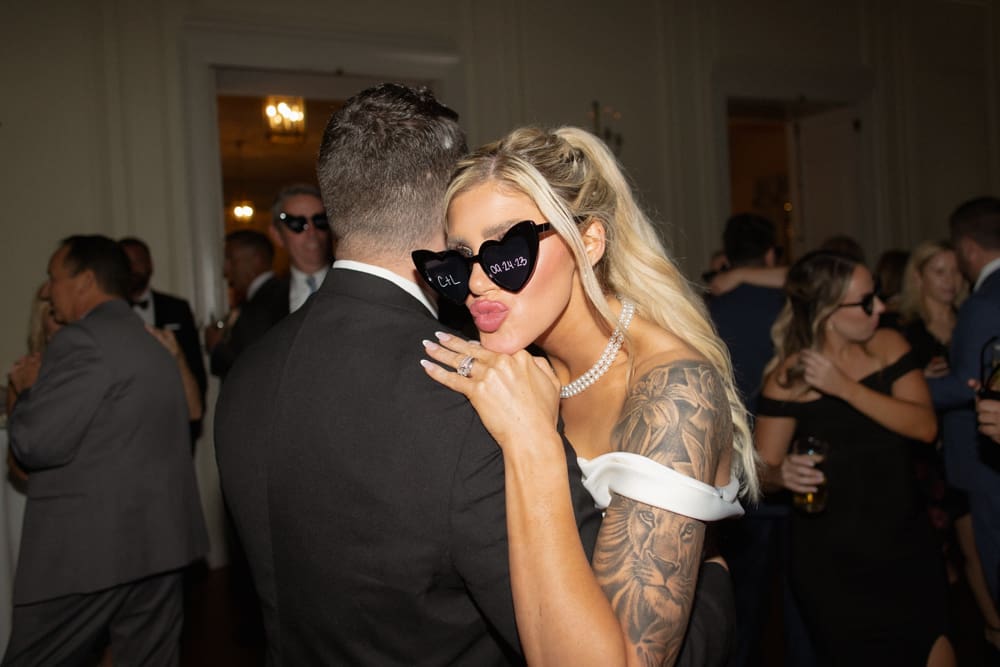Bride wearing heart-shaped sunglasses leaning into the groom on the dance floor during the reception.