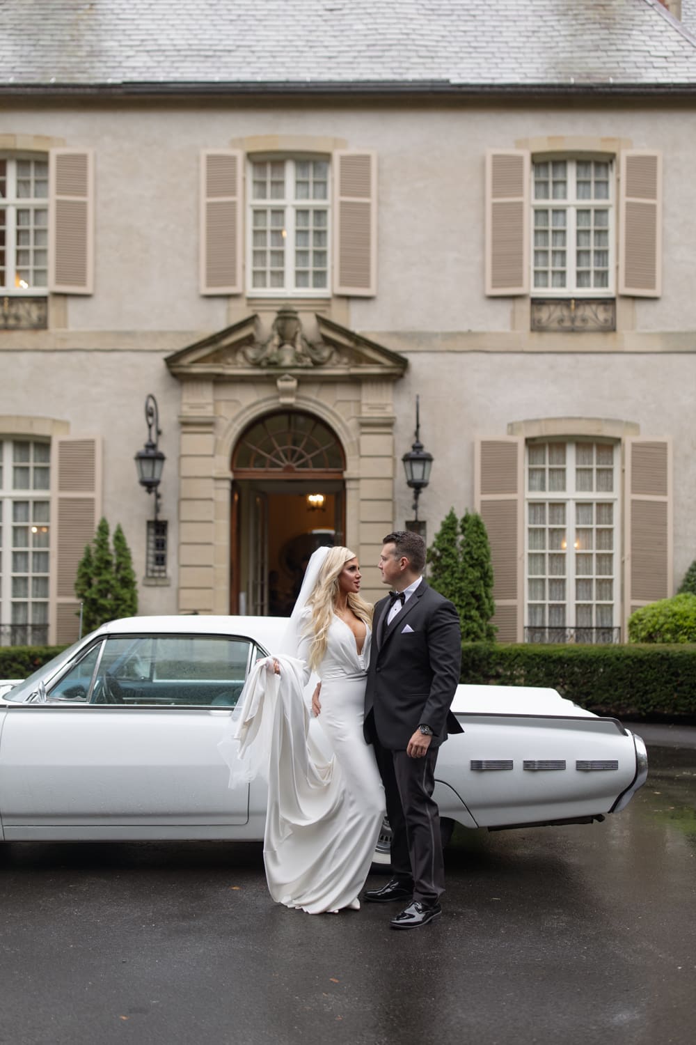Bride and groom standing beside a vintage car outside a historic estate wedding venue in Rhode Island, looking at each other.
