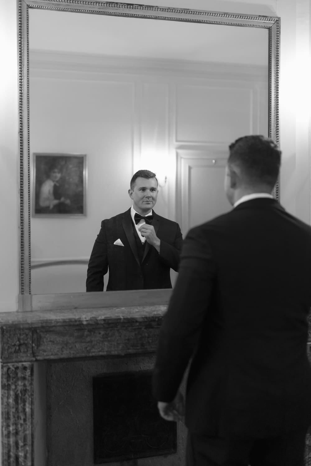 Groom standing in front of a mirror adjusting his bow tie, captured in a quiet moment before the ceremony.
