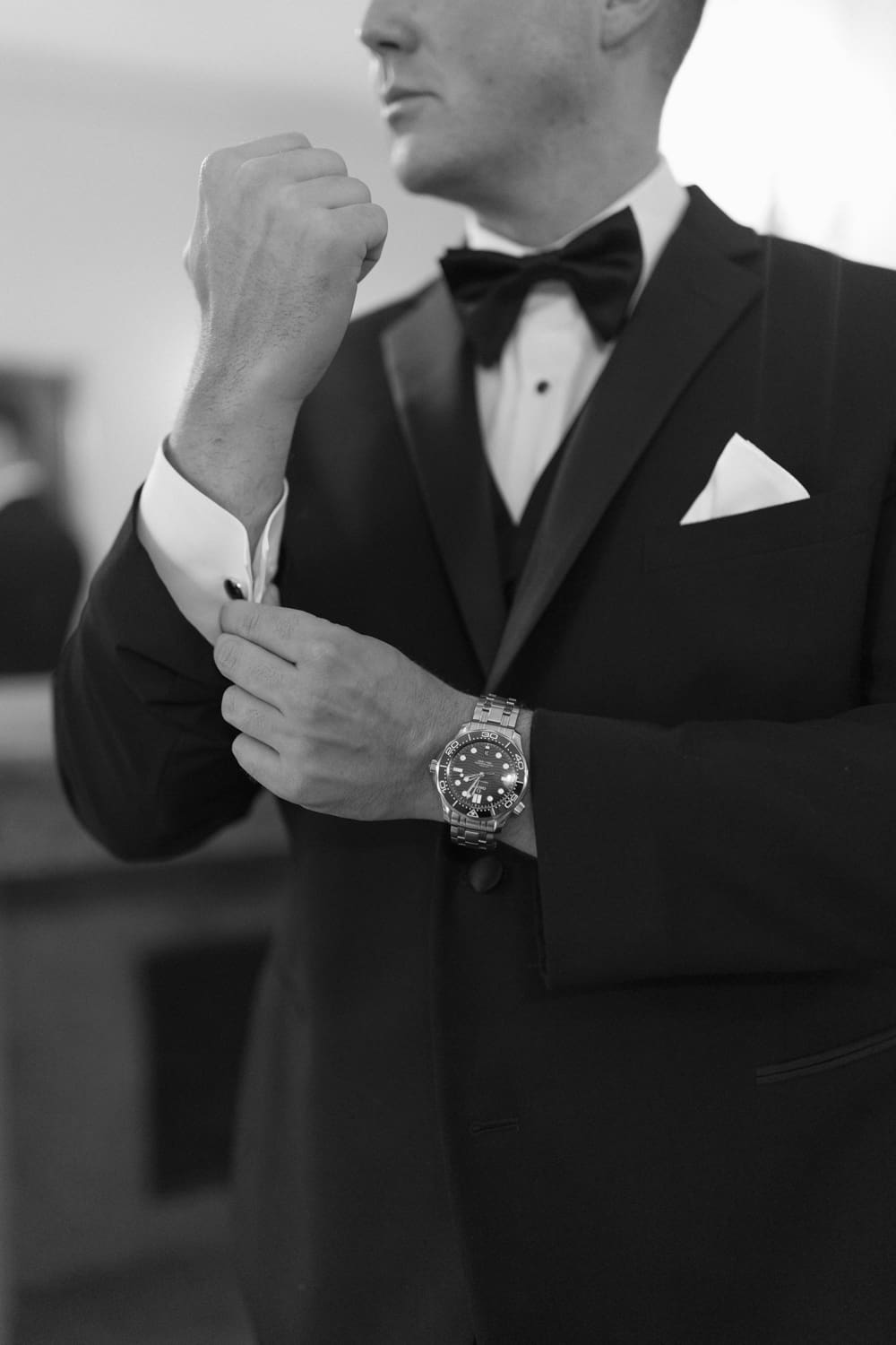 Black and white close-up of groom adjusting his cufflinks, wearing a classic tuxedo and watch.
