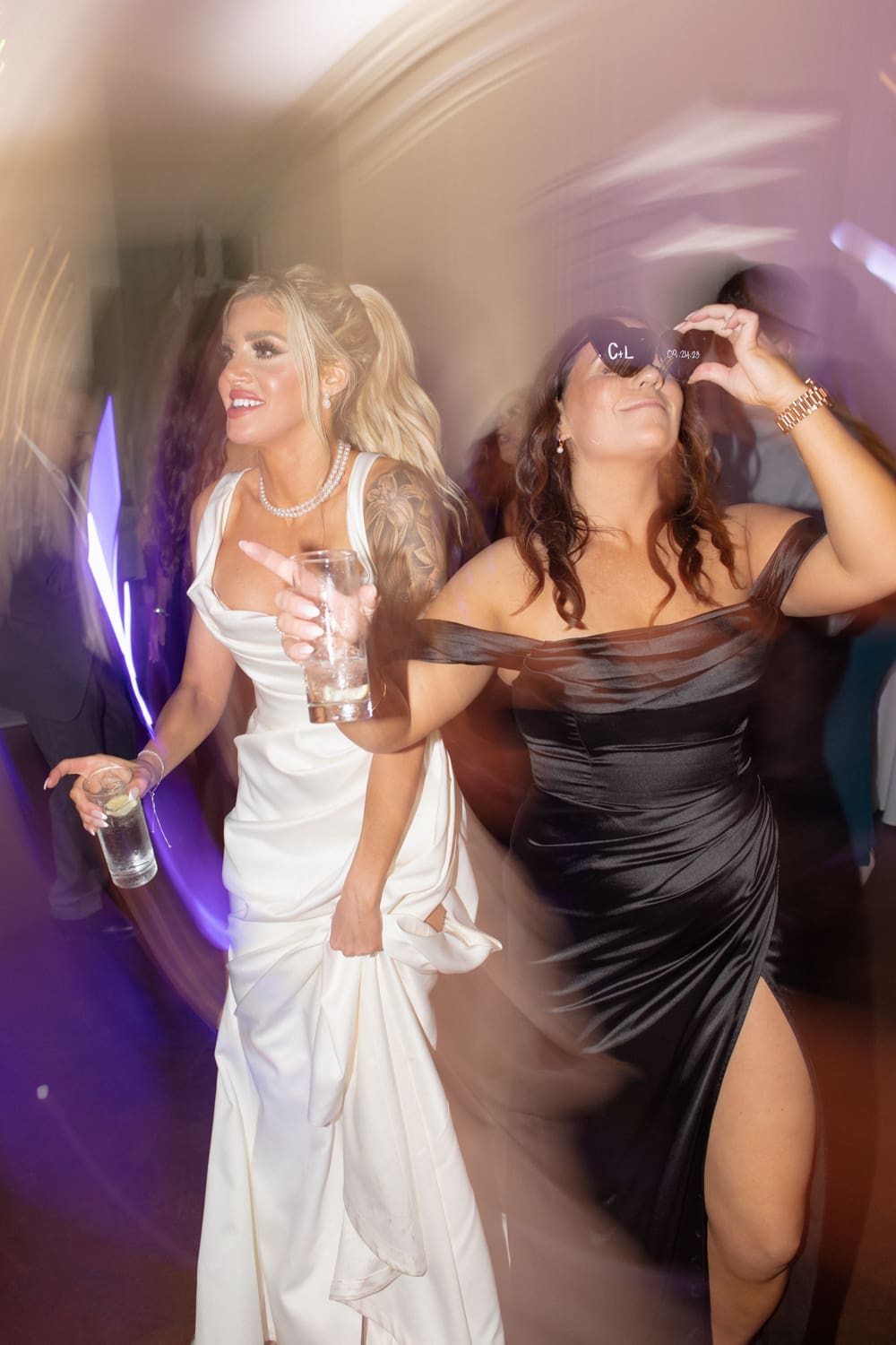 Bride and guest dancing with drinks in hand under colorful reception lighting at a wedding venue in Rhode Island.