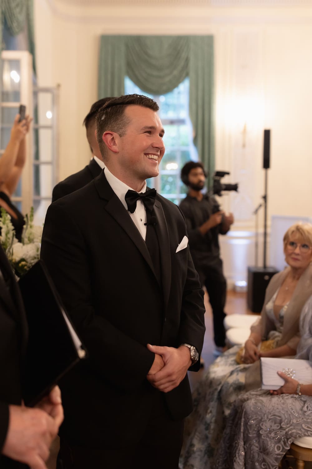 Groom smiling during the ceremony as he looks toward the bride, with guests softly watching in the background.
