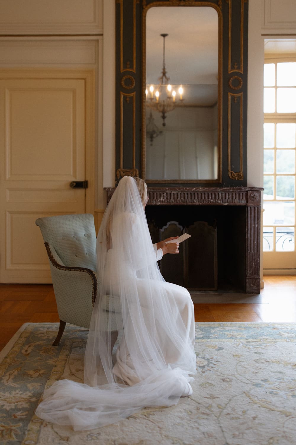 Bride seated in a soft blue chair reading a note, her veil draped across the floor in a calm getting ready space.
