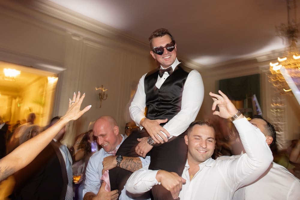 Groom lifted on guests’ shoulders while dancing in a crowded reception ballroom at a wedding venue in Rhode Island.